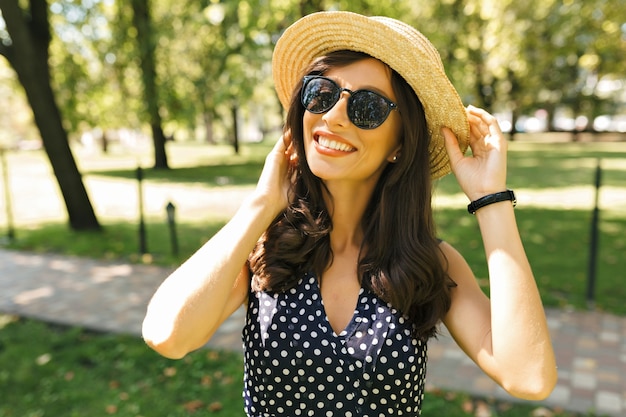 Photo of pretty woman with dark short hair dressed in cute dress with charming smile. She wearing summer hat and black sunglasses. Beautiful portrait.