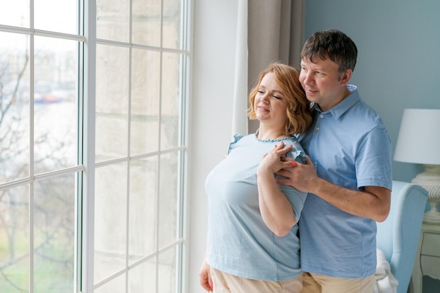 Photo of romantic caucasian cute couple smiling and hugging while standing by the window of a house in a bright bedroom in casual light blue clothes Married couples relationship concept