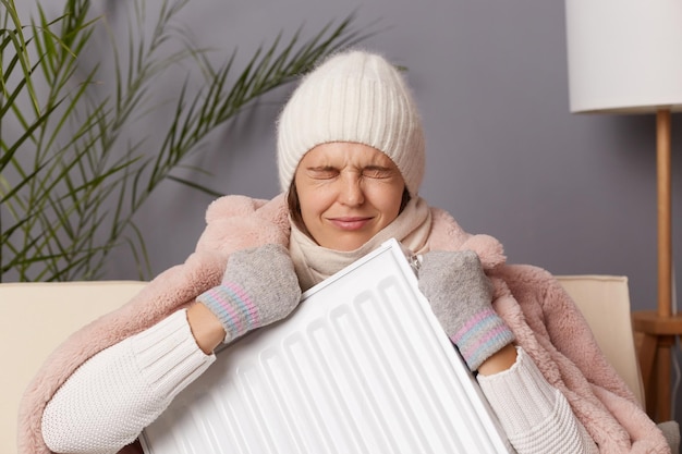 Photo of sad depressed woman in coat and hat sit in cold living room holding in hands radiator struggle from chill freeze at home and crying while sitting in cold house