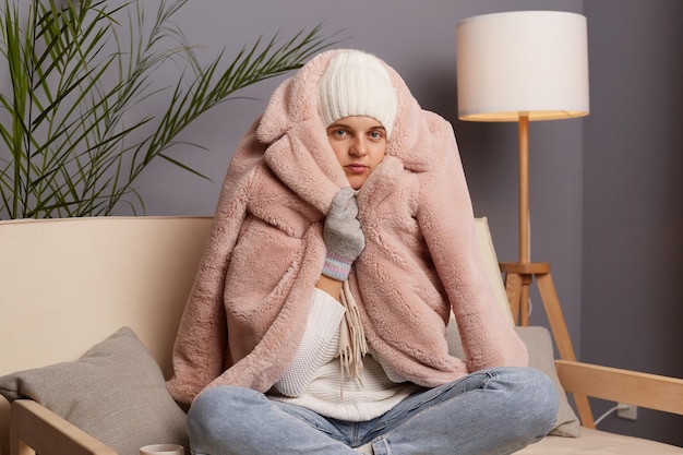 Photo of sad unhappy cold woman wearing warm clothing sitting on sofa in room wrapped in coat posing with crossed legs expressing negative emotions