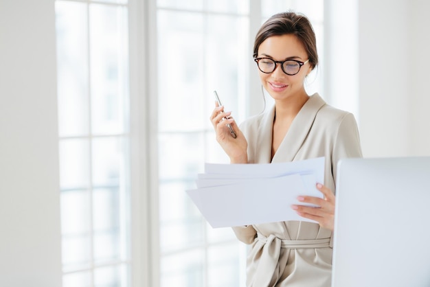 Photo of satisfied young woman with dark hair dressed in business suit focused in papers works in office holds modern cellphone wears optical glasses for good vision has pleased expression