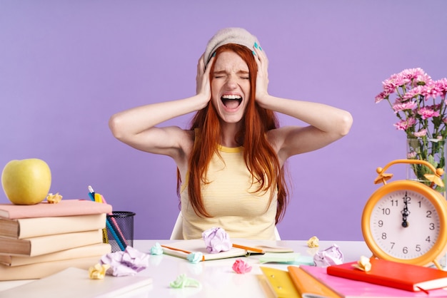 Photo of screaming student girl grabbing her head while sitting at desk with exercise books isolated over purple wall