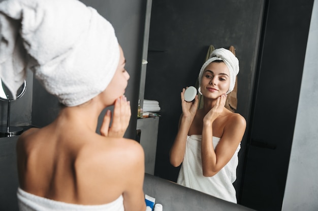 Photo of seductive woman wrapped in towel applying cream at face