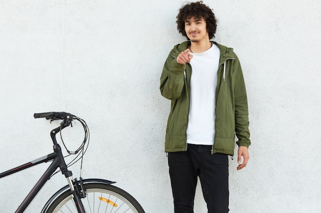 Photo of self confident male has curly hair, dressed in fashionable anorak, points with index finger directly , makes choice