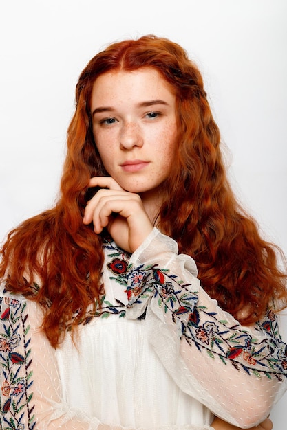 Photo of serious beautiful ginger woman smooth healthy skin has red wavy natural hair dressed in casual white jumper looks at camera poses in studio copy space on right side