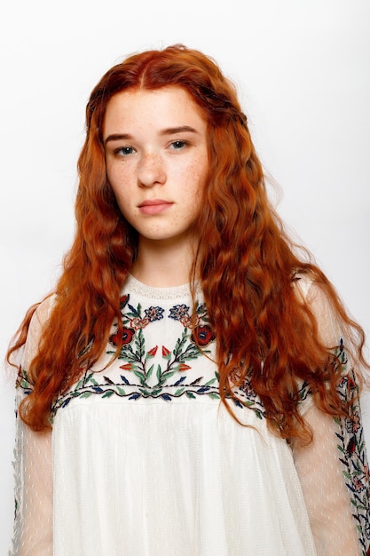 Photo of serious beautiful ginger woman smooth healthy skin has red wavy natural hair dressed in casual white jumper looks at camera poses in studio copy space on right side