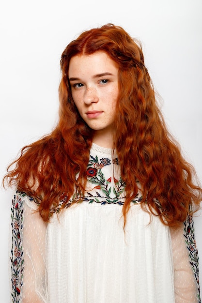 Photo of serious beautiful ginger woman smooth healthy skin has red wavy natural hair dressed in casual white jumper looks at camera poses in studio copy space on right side