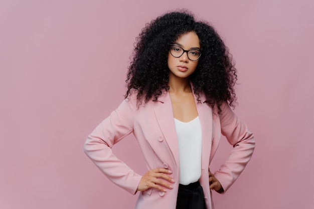 Photo of serious curly haired woman looks confidently at camera wears formal neat clothes keeps both hands on waist wears optical glasses poses over purple background People style concept