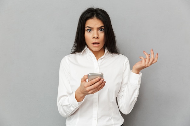 Photo of shocked or outraged woman 30s in formal wear looking on camera while holding smartphone in hand, isolated over gray background