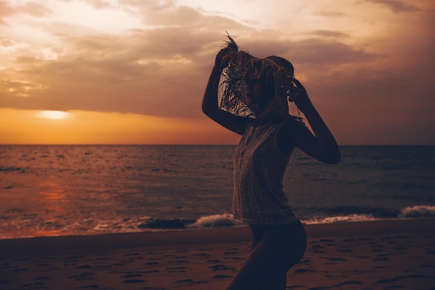 Photo photo shoot at sunset. a nice young woman in a lace top and a straw hat stands by the ocean with her hands raised and looks away. tropical landscape with views of the sea and the coast.