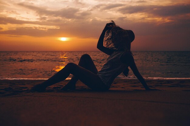 Photo photo shoot at sunset. a young beautiful woman in a straw hat and a lace top elegantly poses sitting on the sand looking to the side holding her hat with her hand. atmospheric tropical landscape.