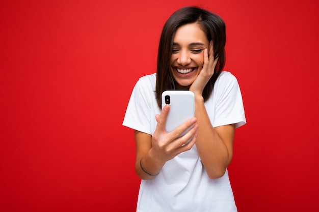 Photo shot of attractive positive surprised good looking young brunette woman wearing casual stylishwhite t-shirt poising isolated on red background with empty space holding in hand and using mobile p