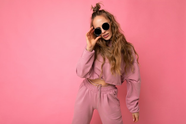 Photo shot of beautiful young dark blonde woman wearing casual clothes and stylish sunglasses isolated over colorful wall .