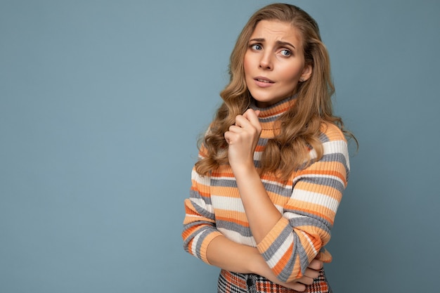 Photo shot portrait of young nice winsome beautiful sad upset thoughtful blonde woman with sincere emotions wearing striped sweater isolated over blue wall with free space and worried about.