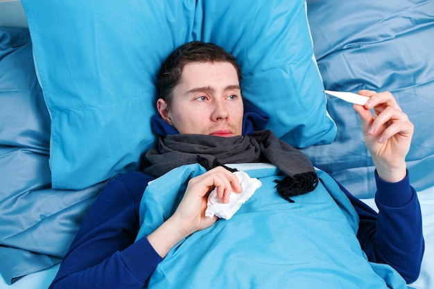 Photo of sick man in scarf with thermometer in hand lying in bed