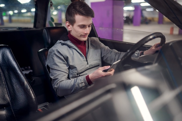 Photo of side of young male driver with phone in hands sitting in car