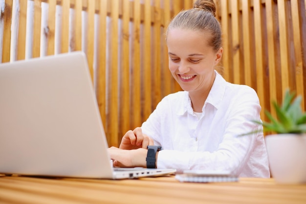 Photo of smiling delighted attractive woman with hair bun wearing white shirt sitting and working on laptop checking her smartwatch with positive facial expression