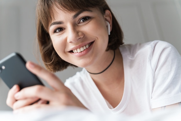 Photo of a smiling pleased young beautiful woman lies indoors at home on bed listening music with bluetooth earphones using mobile phone.