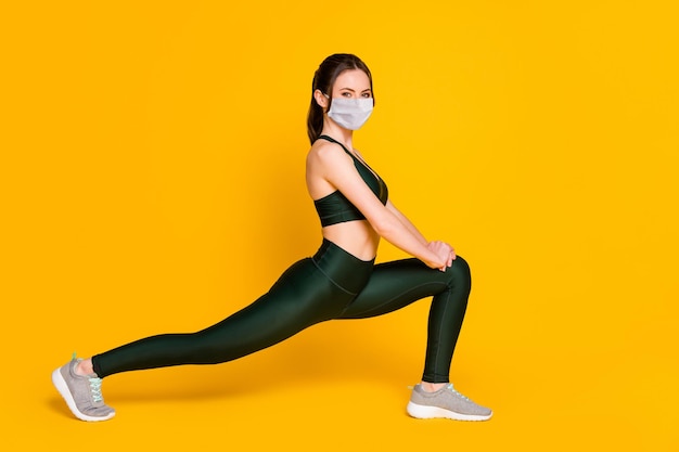 Photo of sportive young lady stretching sit one leg wear mask sports suit top pants sneakers isolated yellow color background