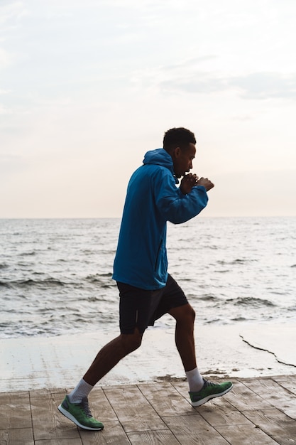 Photo of a strong handsome young african sports man boxer outdoors at the beach sea walking make boxing exercises.