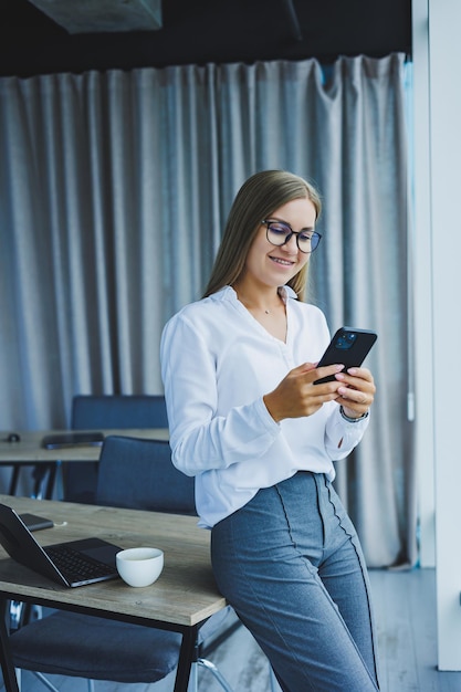 Photo of a successful young woman in a shirt smiling and working on a laptop while talking on the phone in a modern office with large windows Remote work with coffee