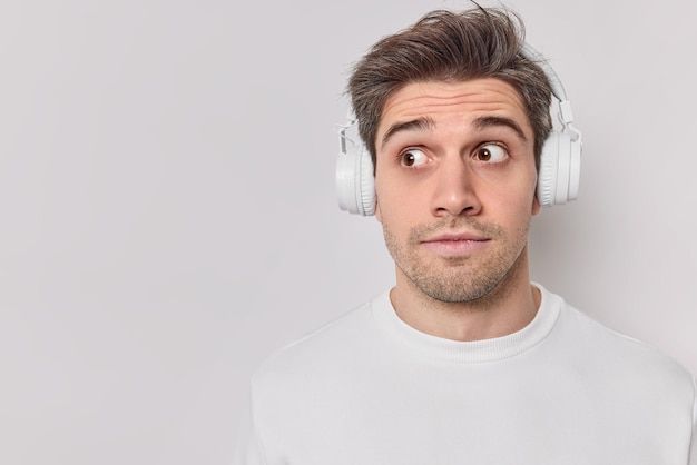 Photo of surprised unshaven guy concentrated away with shocked expression has widely opened eyes listens audio track in wireless headphones wears jumper isolated over white background copy space