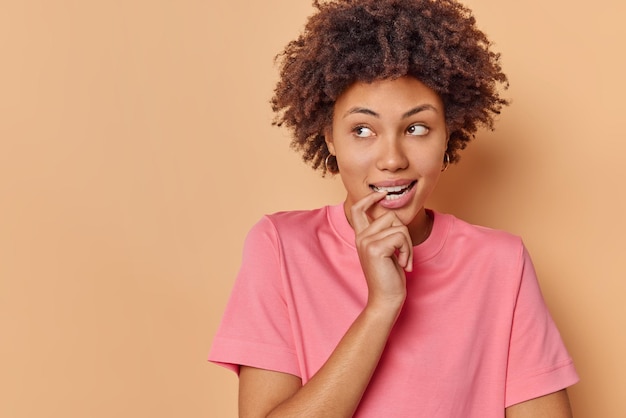 Photo of thoughtful dreamy young woman bites finger calculates in mind makes decision ponders on something wears casual pink t shirt isolated over beige background copy space for your promotion