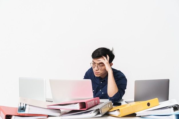 Photo of tired asian man 30s wearing eyeglasses sitting at table and using laptops while working in bright office room