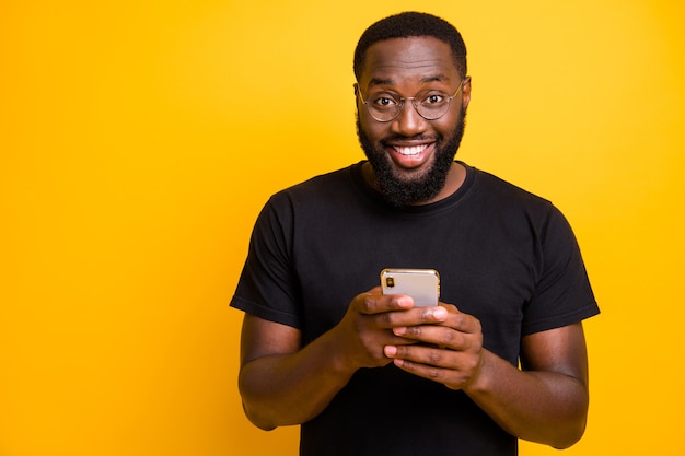 Photo of trendy cheerful positive handsome man smiling toothily in spectacles browsing through his telephone in t-shirt isolated vivid color wall 