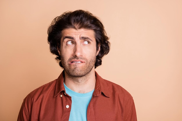 Photo of uncertain bearded guy bite lip look empty space wear brown t-shirt isolated beige color background