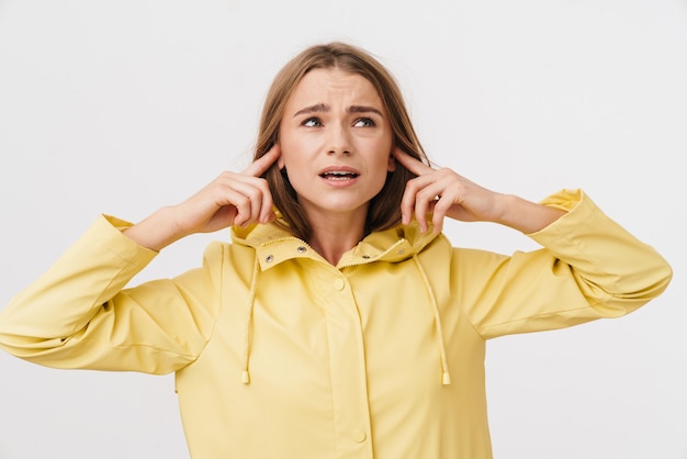 Photo of unhappy young woman in raincoat plugging her ears and looking upward 