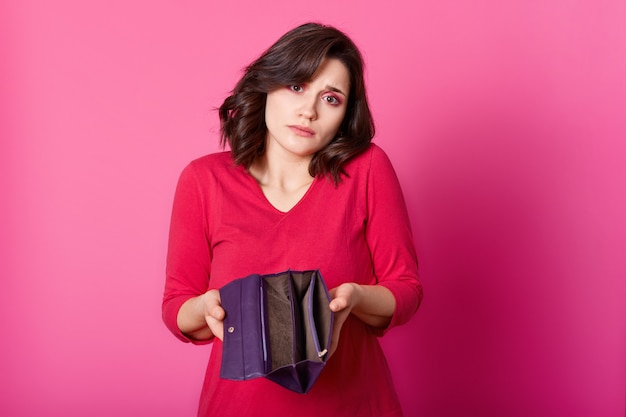 Photo of upset brunette with opened wallet in hands. Beautiful sad woman squeezes shoulders and does not khow how to pay for purchases. Attractive girl wears red jumper stands against pink wall.