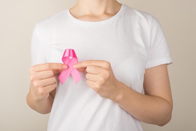 Photo of woman in white tshirt holding pink ribbon symbol of breast cancer awareness on isolated grey background