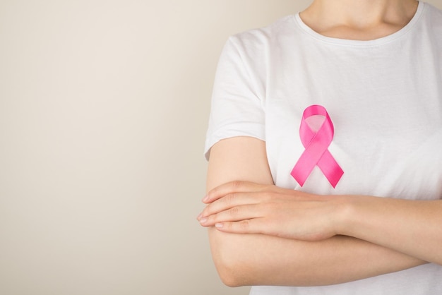 Photo of woman with crossed arms in white tshirt with pink ribbon symbol of breast cancer awareness on isolated grey background with copyspace