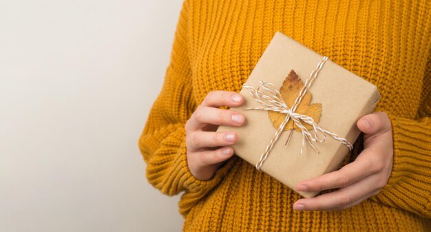 Photo of woman in yellow knitted sweater holding craft paper giftbox with twine bow and orange autumn leaf on isolated white background