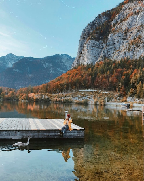 Photo of woman in yellow raincoat sitting on the bridge on the lake at autumn mountains landscape