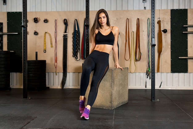 Photo workout of a beautiful brunette girl in a black sports uniform and pink sneakers which sits on a wooden cube in the gym