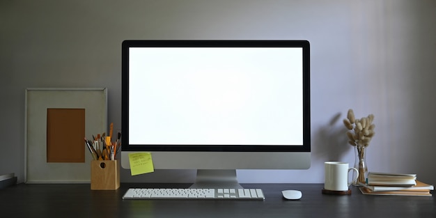 Photo of Workspace blank screen computer monitor putting on working desk and surrounded by picture frame, pencil holder, stack of books, wireless mouse, keyboard, coffee cup and wild grass in vase.