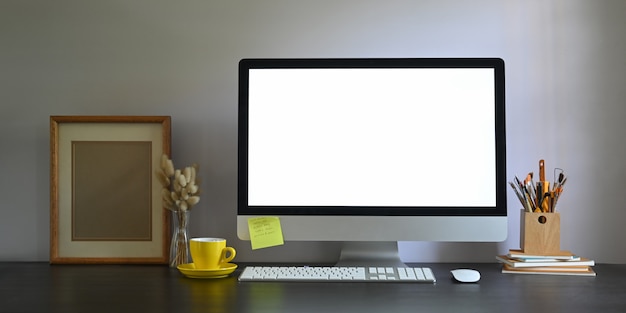 Photo of Workspace blank screen computer monitor putting on working desk and surrounded by picture frame, pencil holder, stack of books, wireless mouse, keyboard, coffee cup and wild grass in vase.