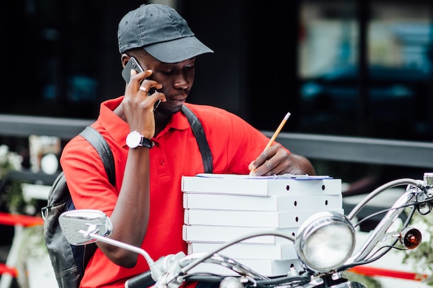 Photo of young african guy accepts the order by phone and write in motorbike holding boxes with pizza. Urban place.