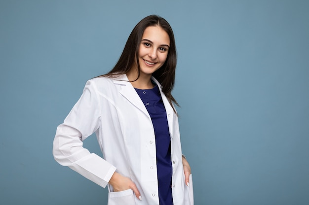 Photo of young attractive beautiful positive smiling brunette woman with sincere emotions wearing white medical coat isolated over blue wall with empty space.