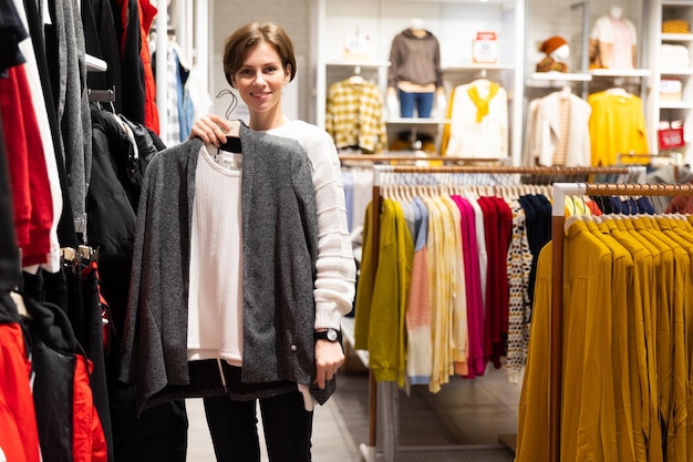 Photo of young attractive brunette woman with a short haircut in a white sweater chooses stylish and casual clothes in a store in a shopping mall. Shop concept.