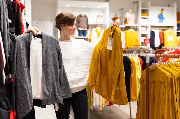 Photo of young attractive brunette woman with a short haircut in a white sweater chooses stylish and casual clothes in a store in a shopping mall. Shop concept.