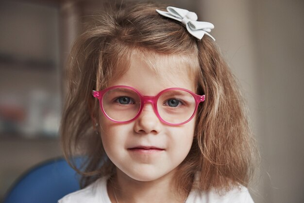 Photo of young blonde haired female child in pink glasses sitting in doctor cabinet.