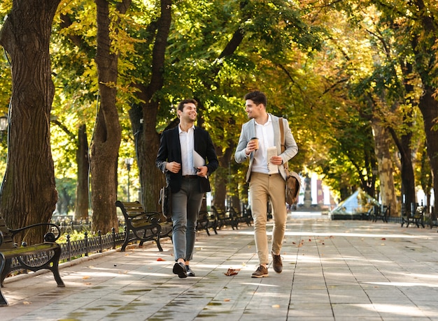 Photo of young businessmen in suits walking outdoor through green park with takeaway coffee and laptop, during sunny day