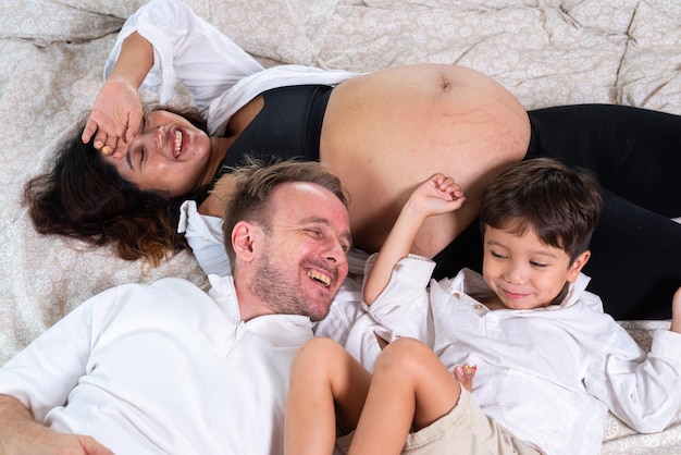 Photo of young cheerful positive smiling couple pregnant wife and husband with their son relaxing at bed