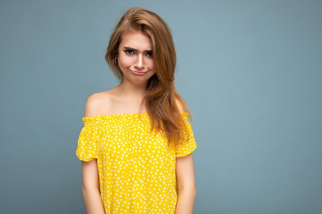 Photo of young cute delightful beautiful attractive funny unhappy dark blonde woman with sincere emotions isolated on blue background wall with copy space for text wearing stylish yellow dress.