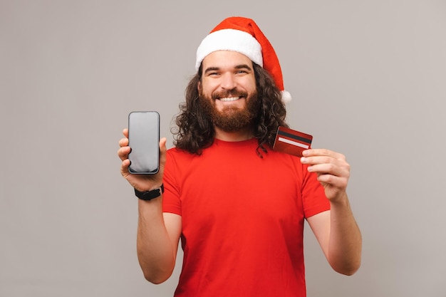 Photo of young happy bearded man wearing santa claus hat and showing credit card and smartphone screen