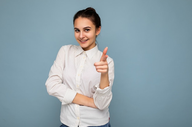 Photo of young happy positive attractive brunette woman with sincere emotions wearing white shirt isolated over blue background with empty space and laughing and pointing at camera with finger