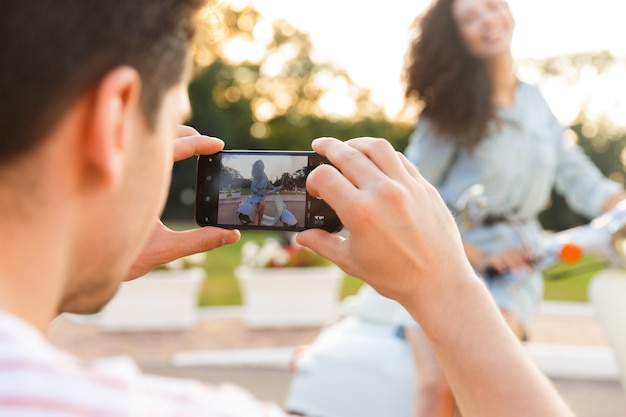 Photo of young man photographing his girlfriend on mobile phone, while she sitting on motorbike in city park